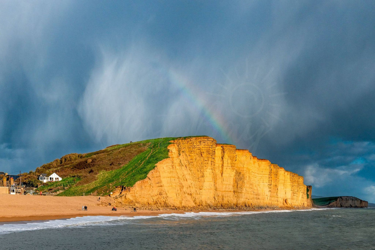 Rainbow above sunlit East Cliff at West Bay, Dorset beneath dark storm clouds