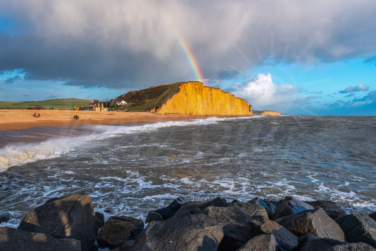 Rainbow over sunlit East Cliff at West Bay Dorset with waves breaking across the shore
