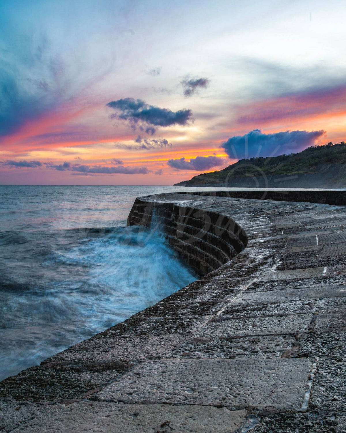 The Cobb at Lyme Regis at sunset, with waves breaking against the stone harbour wall.