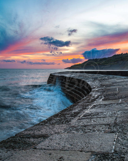 The Cobb at Lyme Regis at sunset, with waves breaking against the stone harbour wall.