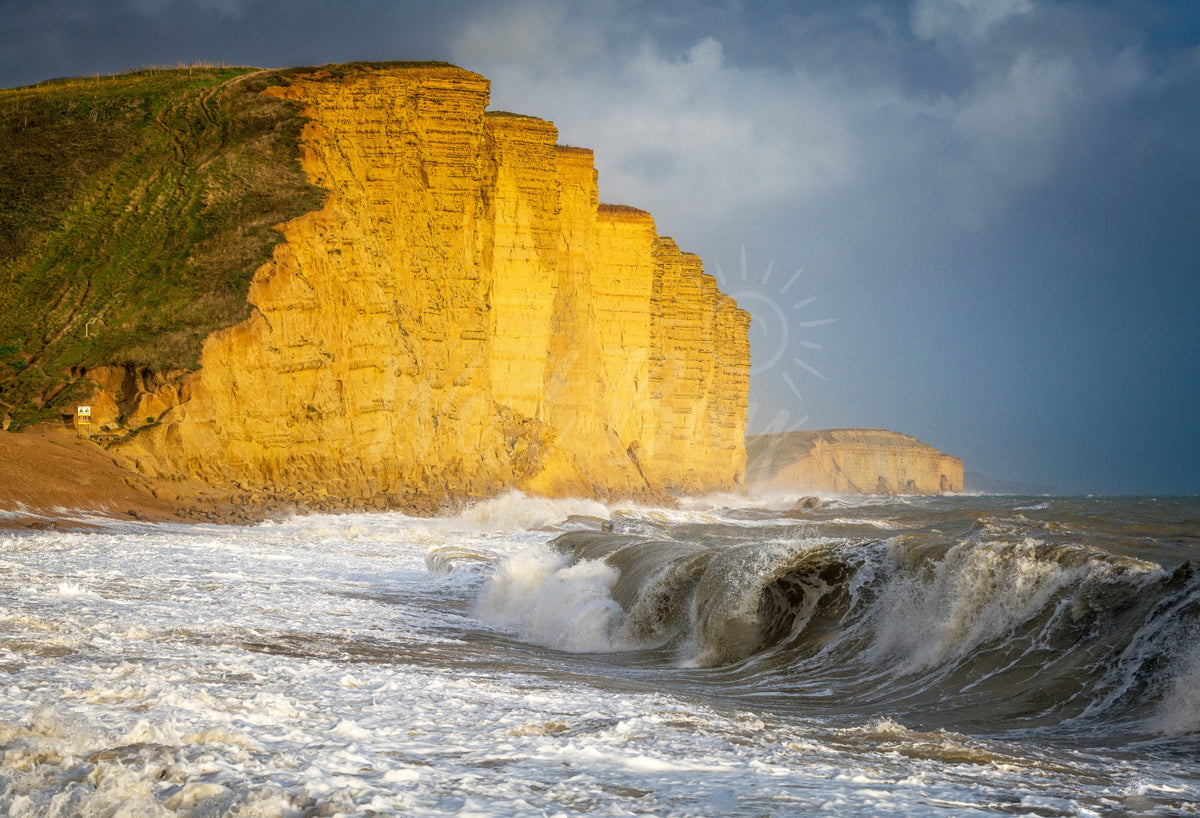 A breaking foreground wave beneath sunlit East Cliff and storm clouds at West Bay, Dorset