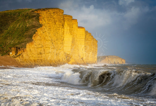 A breaking foreground wave beneath sunlit East Cliff and storm clouds at West Bay, Dorset